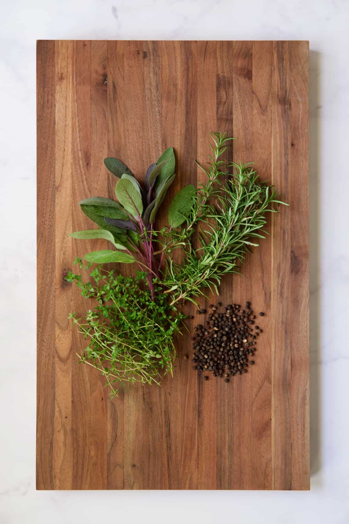 Rosemary, thyme, sage and black peppercorns on wood cutting board.