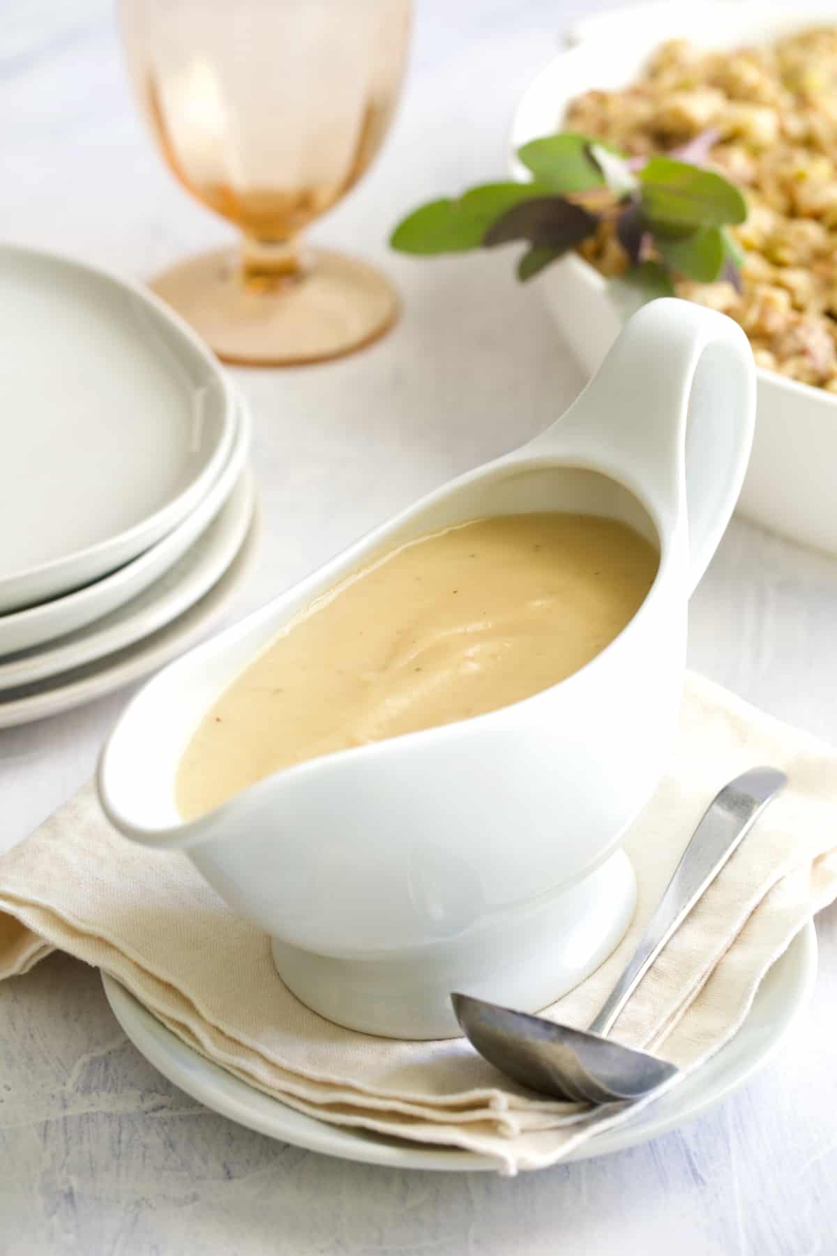 Gravy in white gravy boat on beige napkin with plates, wine glass and stuffing in the background.