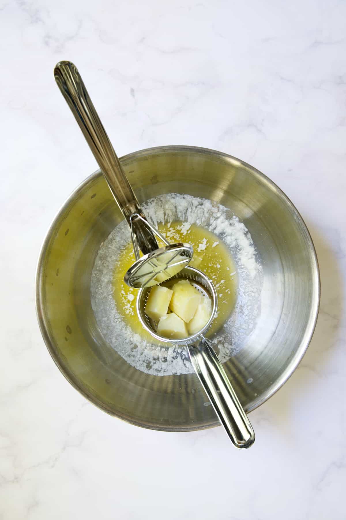 Potatoes in ricer about to be riced into butter in stainless steel bowl.