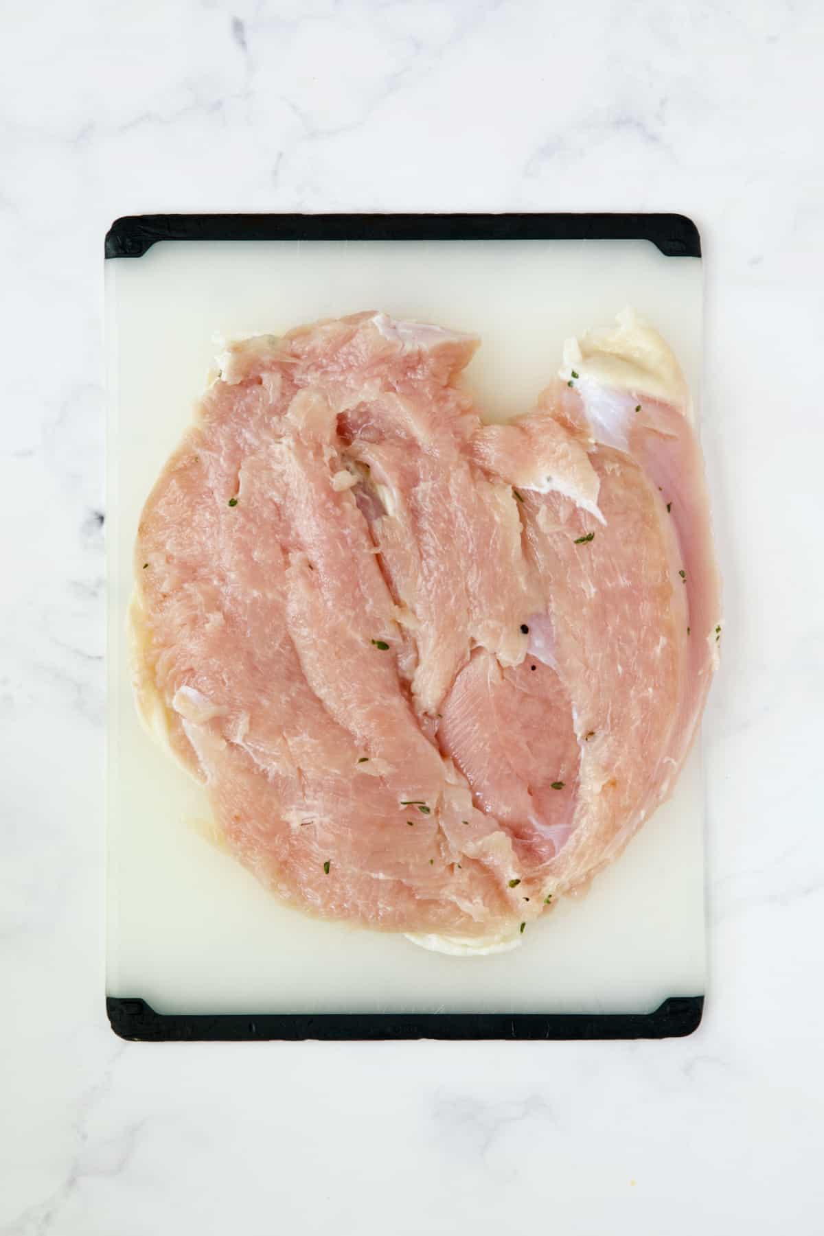 Uncooked flattened turkey breast on white plastic cutting board.
