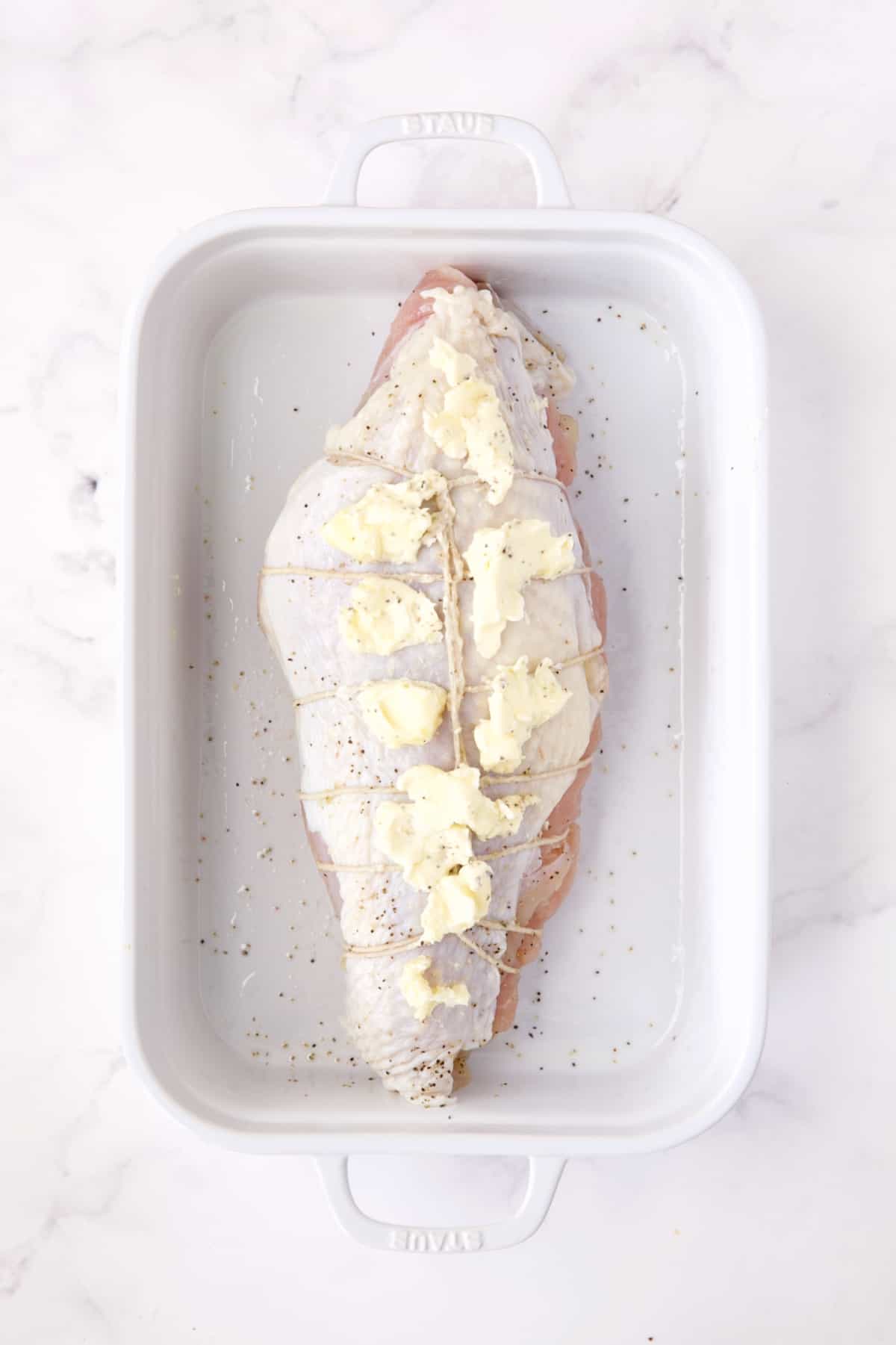 Uncooked stuffed turkey breast in white roasting pan with butter smeared over the skin.