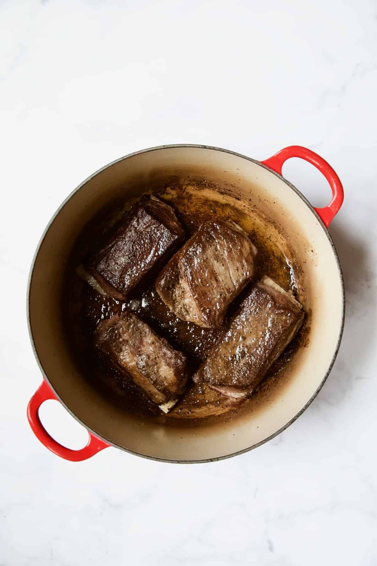 Four beef short ribs after being browned in red enamel Dutch oven.