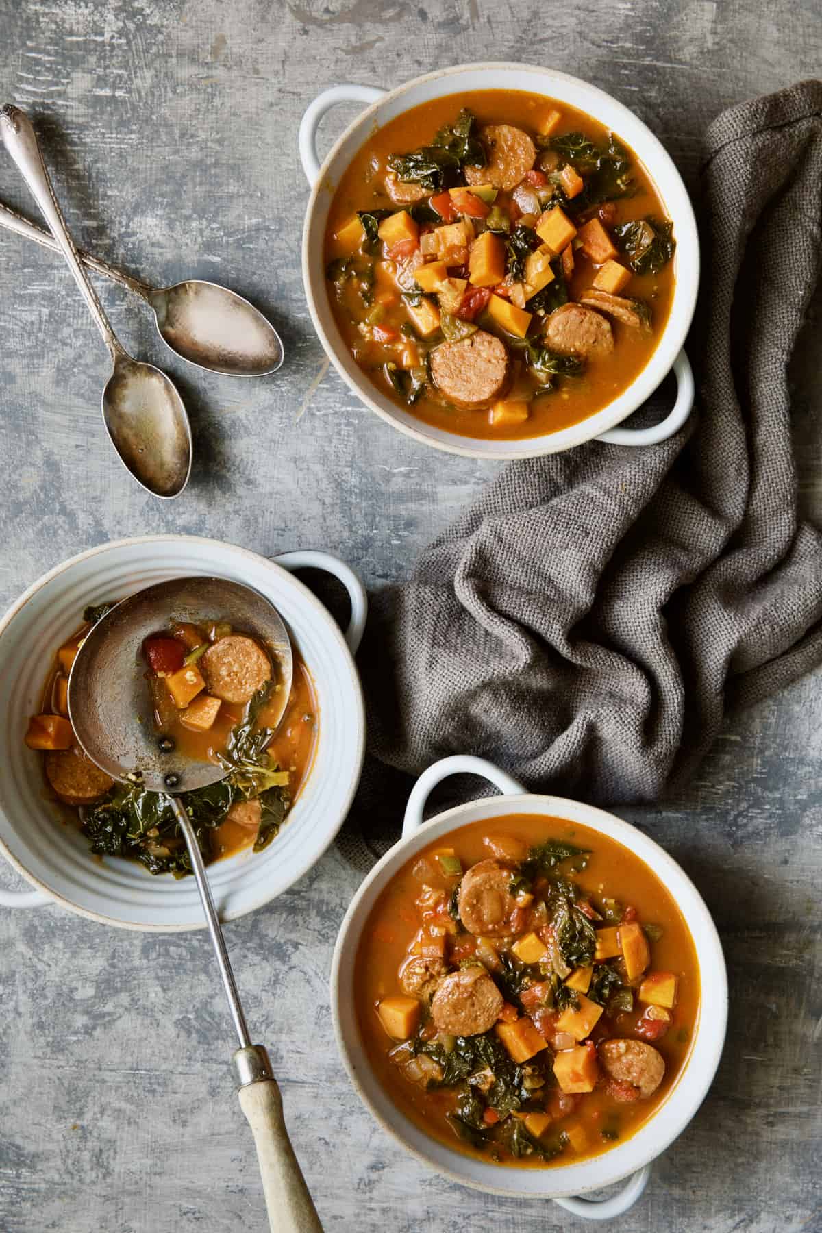 Three bowls of Chorizo Sweet Potato Stew with Kale on gray background with soup spoons.