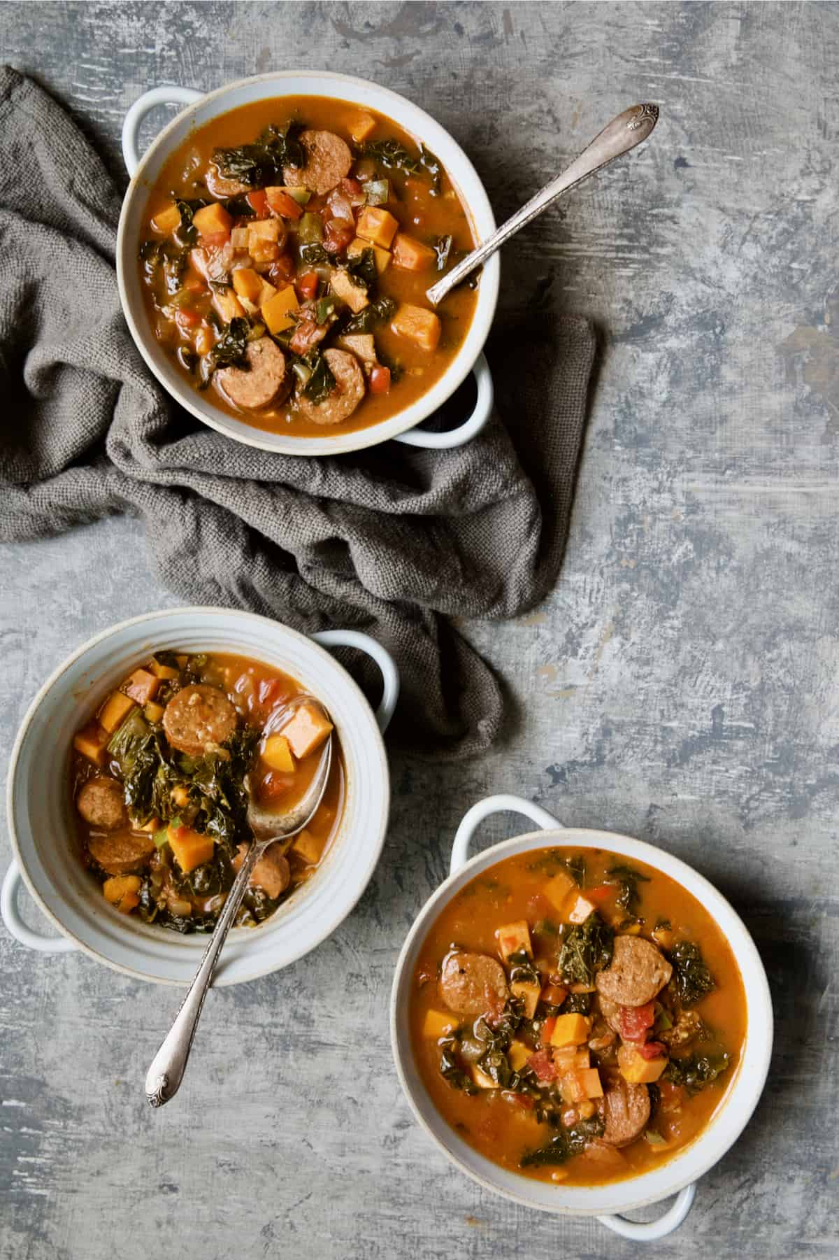 Three bowls of Chorizo Sweet Potato Stew with Kale on gray background with gray napkin.