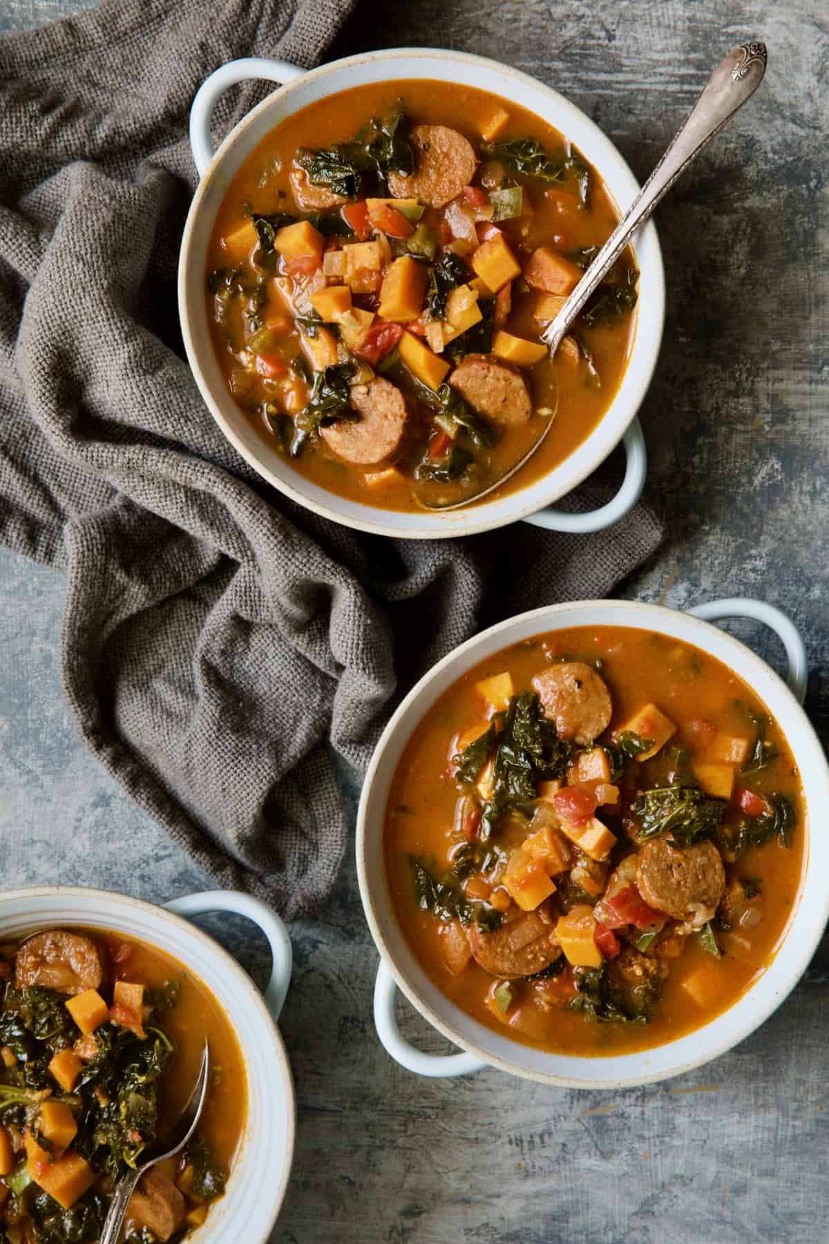 Chorizo Sweet Potato Stew with Kale in white bowls with soup spoon on gray background.