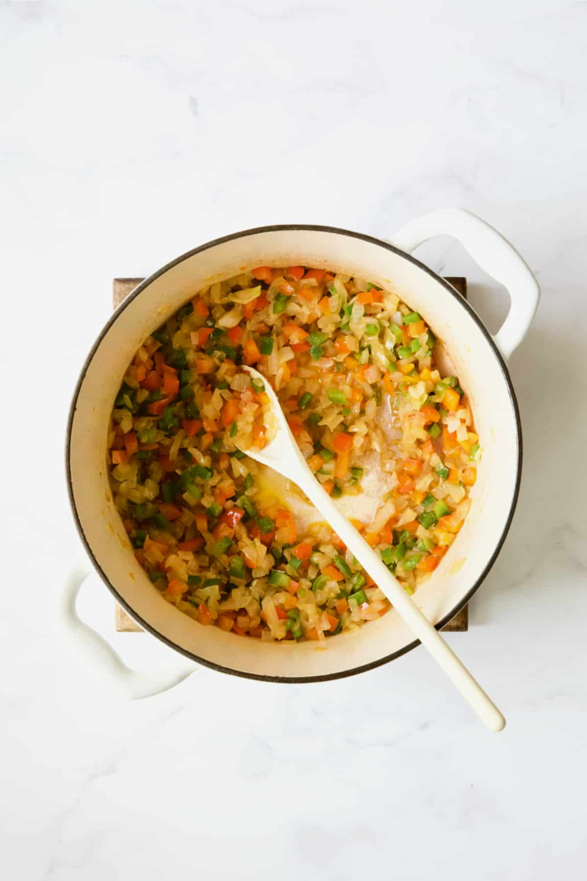 Onions and peppers being cooked in white Dutch oven with white mixing spoon.