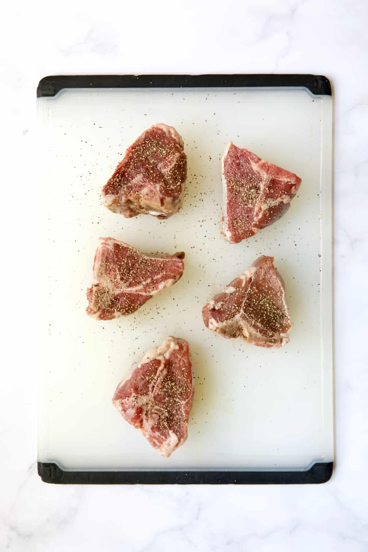 Five lamb chops on white plastic cutting board seasoned with salt and pepper.
