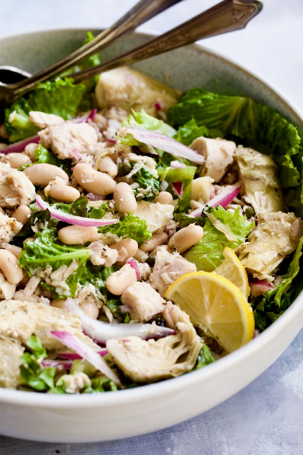 Tuna, White Bean and Artichoke Salad in white serving bowl with silver serving utensils.