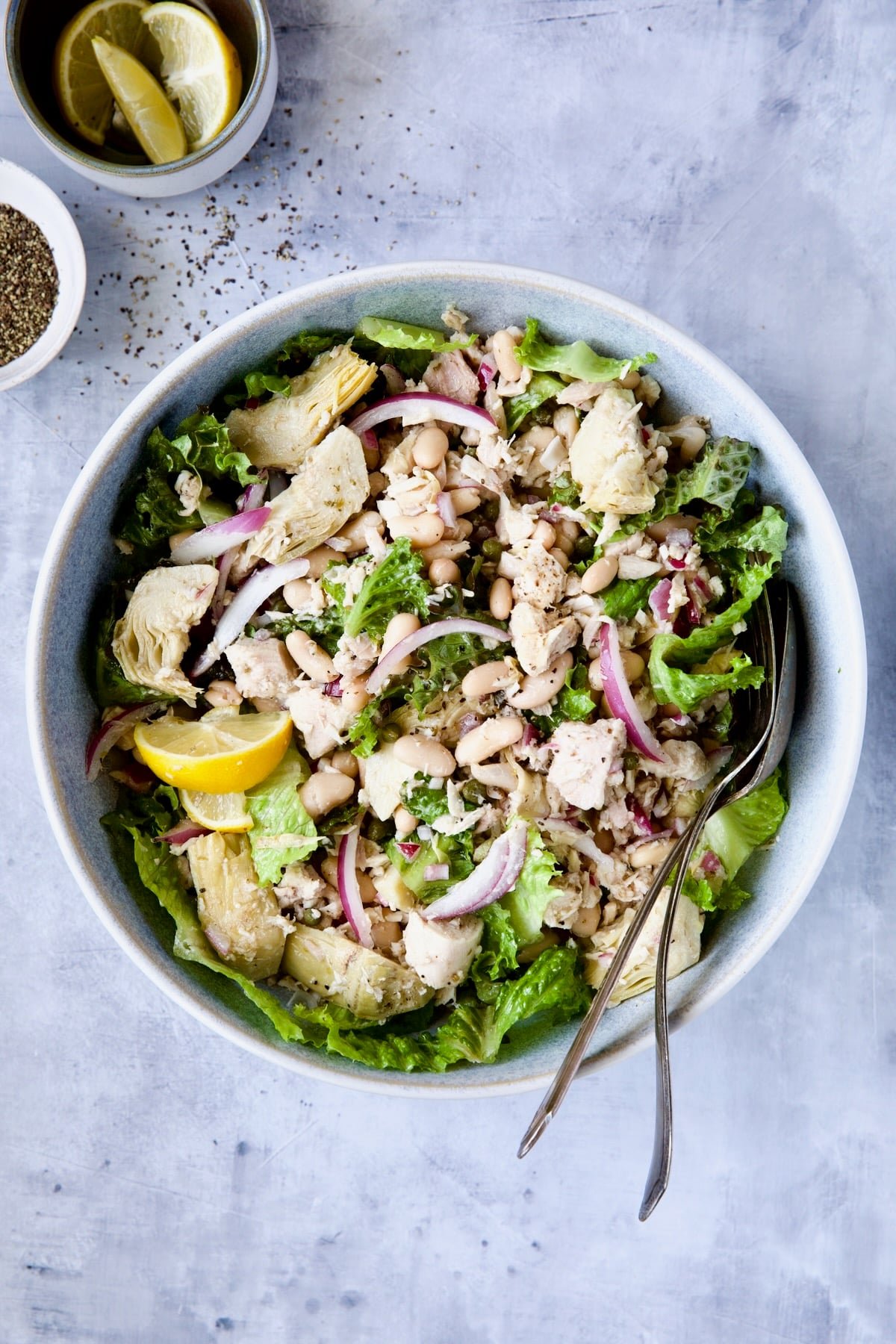 Tuna, White Bean and Artichoke Salad in serving bowl with serving utensils on blue background.