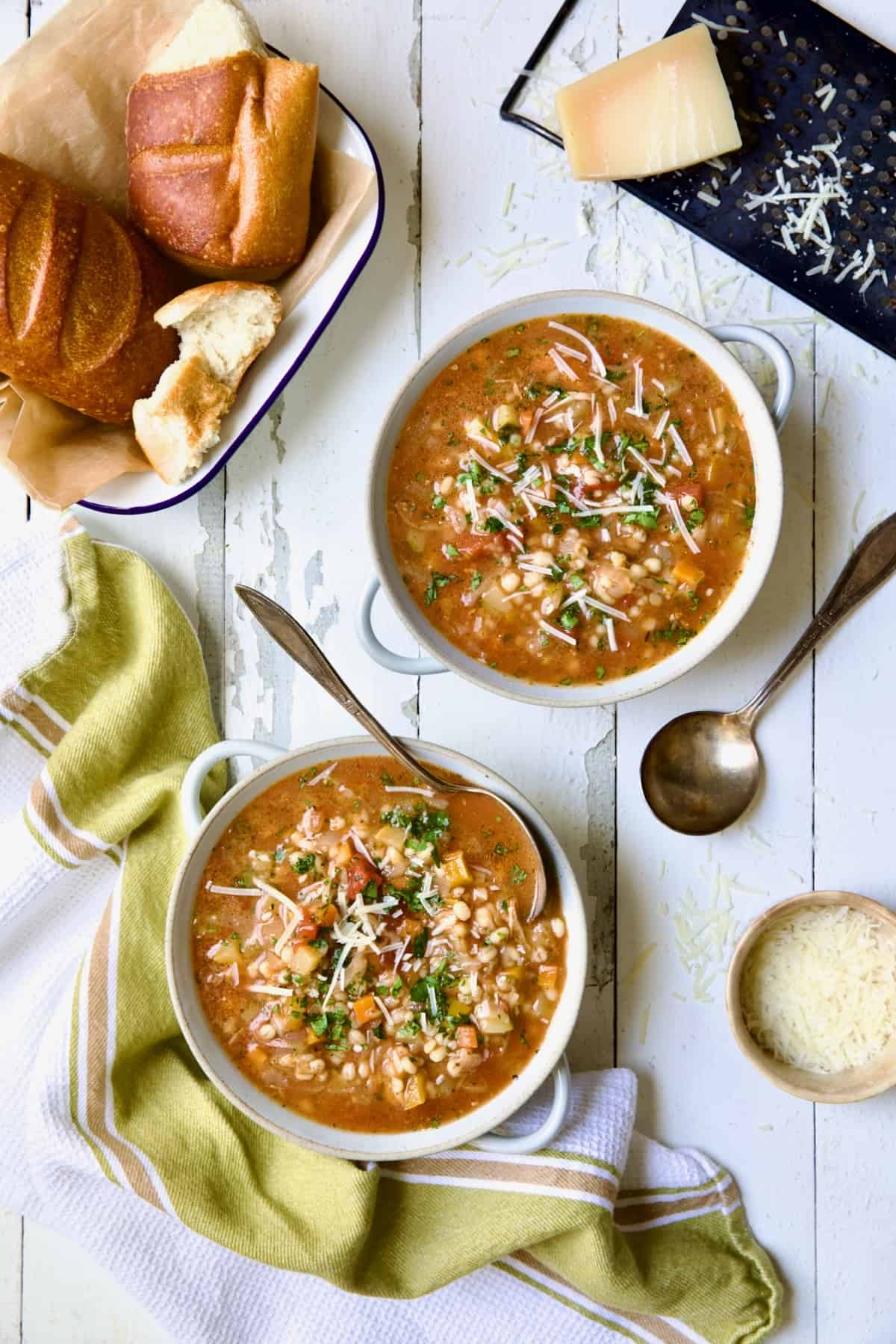 Two bowls of Vegetable Barley Soup on white distressed background with bread and cheese.
