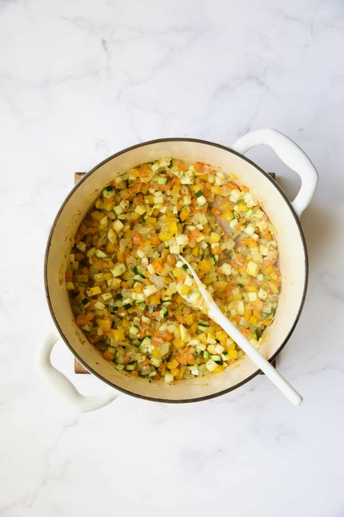 Cooked diced vegetables in white Dutch oven with white mixing spoon.