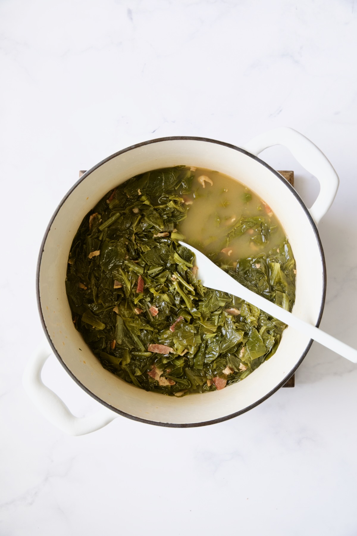 Greens in white Dutch oven being stirred with white spoon.