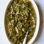 Braised Southern Greens in oval serving bowl with serving spoon on light blue background.