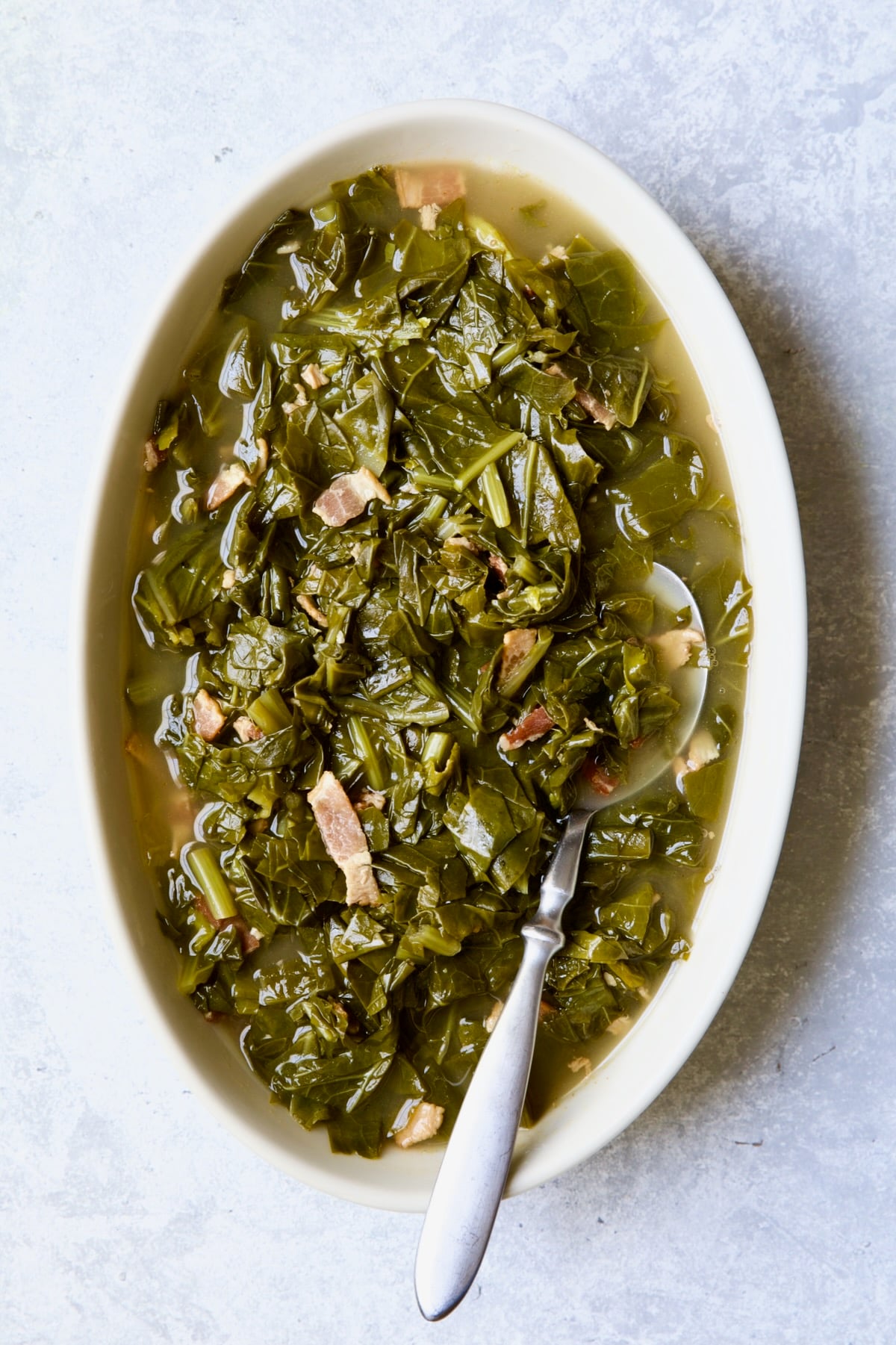 Braised Southern Greens in oval serving bowl with serving spoon on light blue background.
