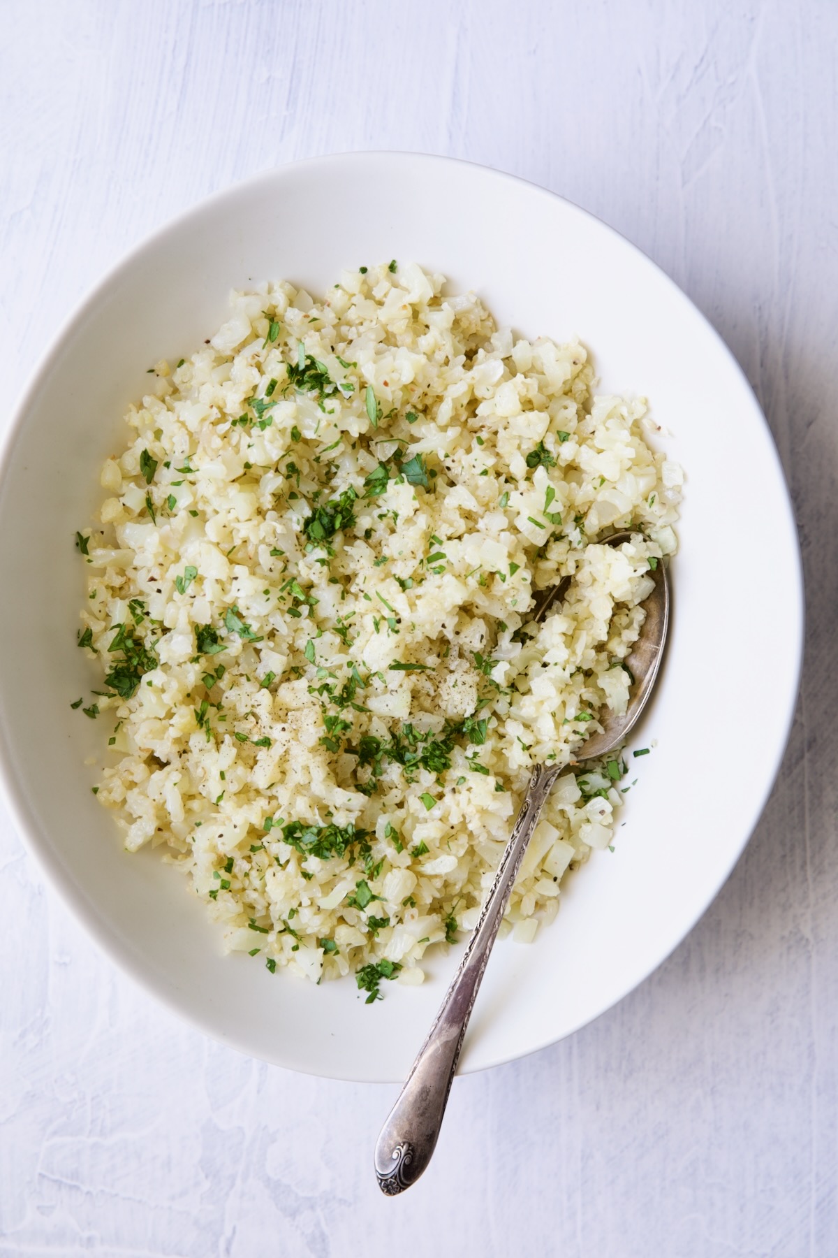 Bowl of plain cauliflower rice in white oval bowl with serving spoon.