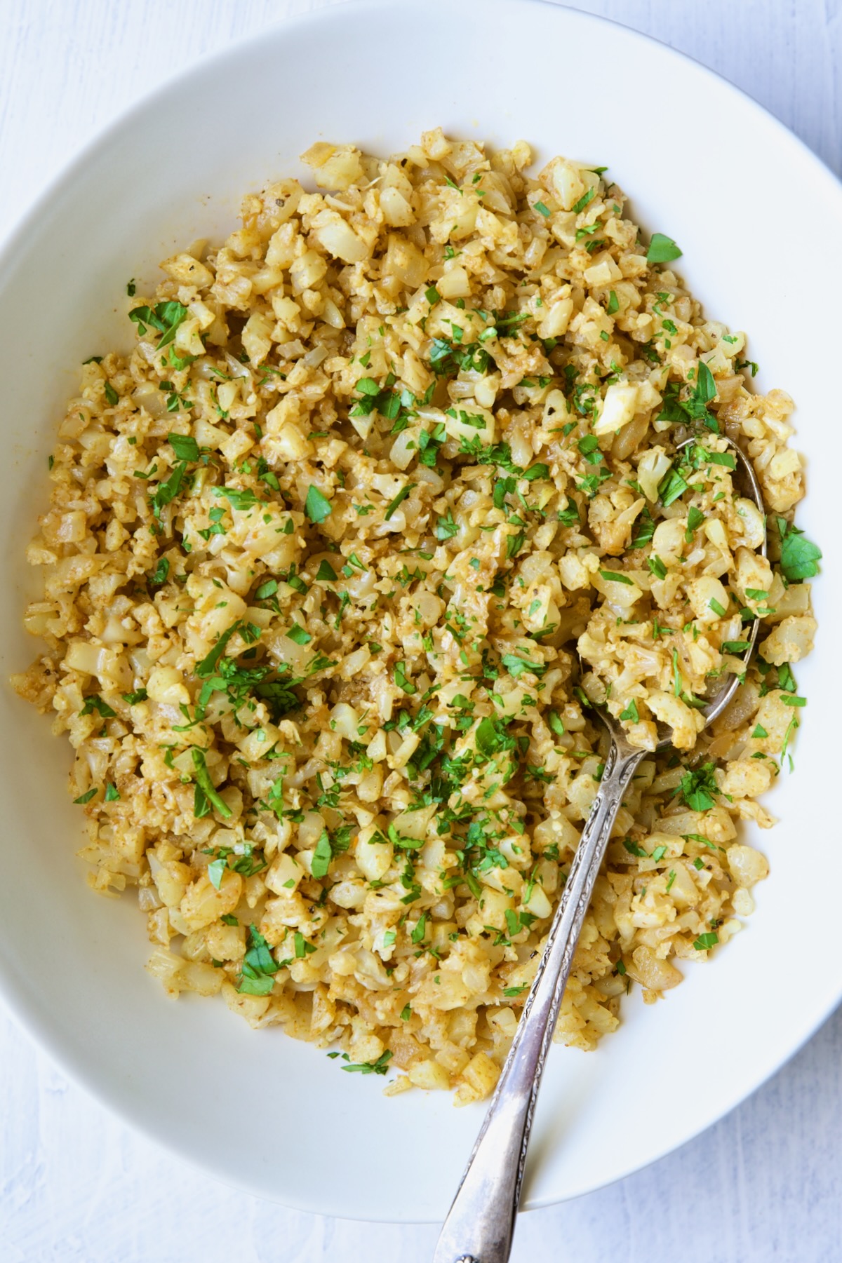 Seasoned cauliflower rice in white oval bowl with serving spoon.