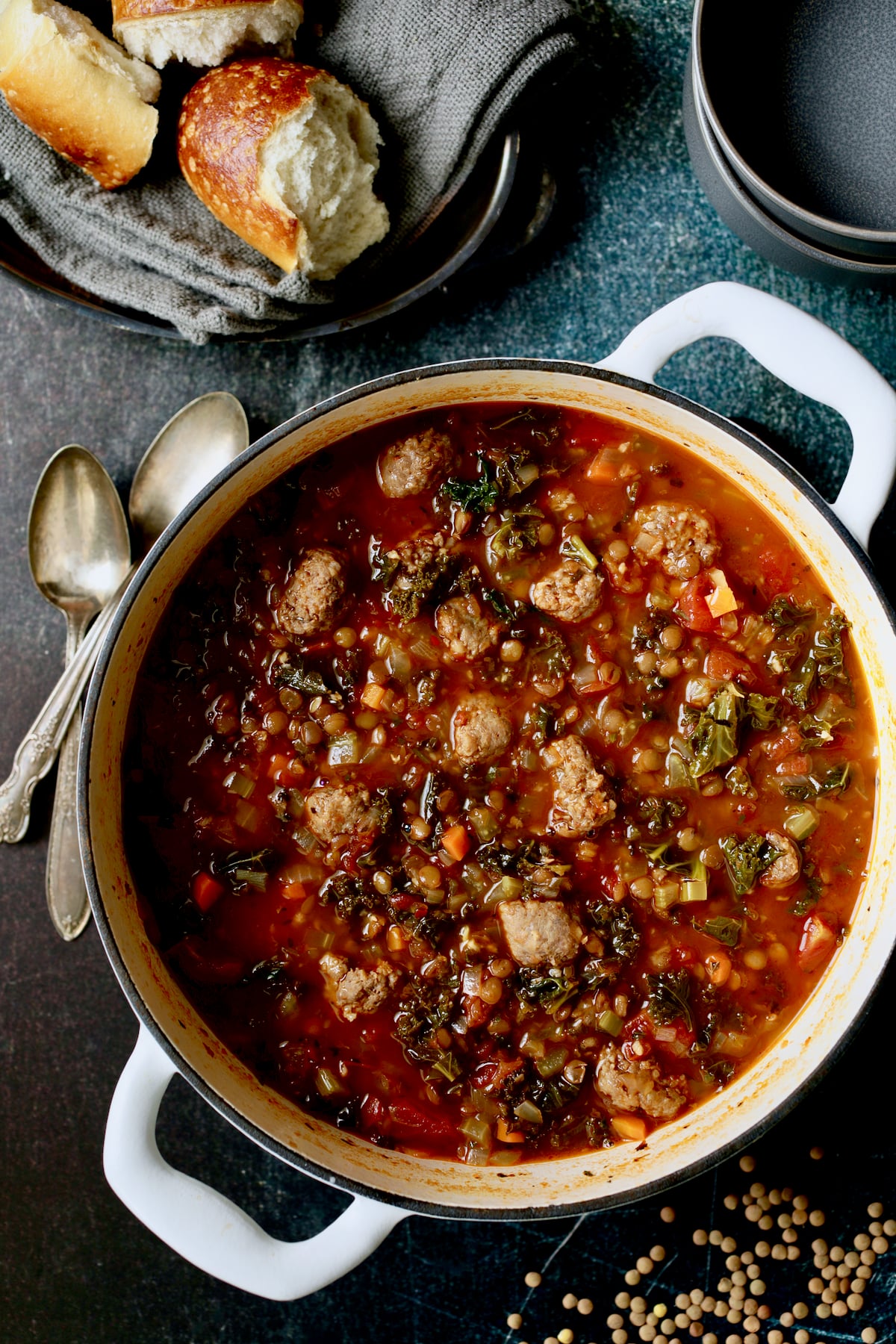 Sausage and Lentil Soup with Kale in white Dutch oven, two spoons, two gray bowls and bread on gray napkin.