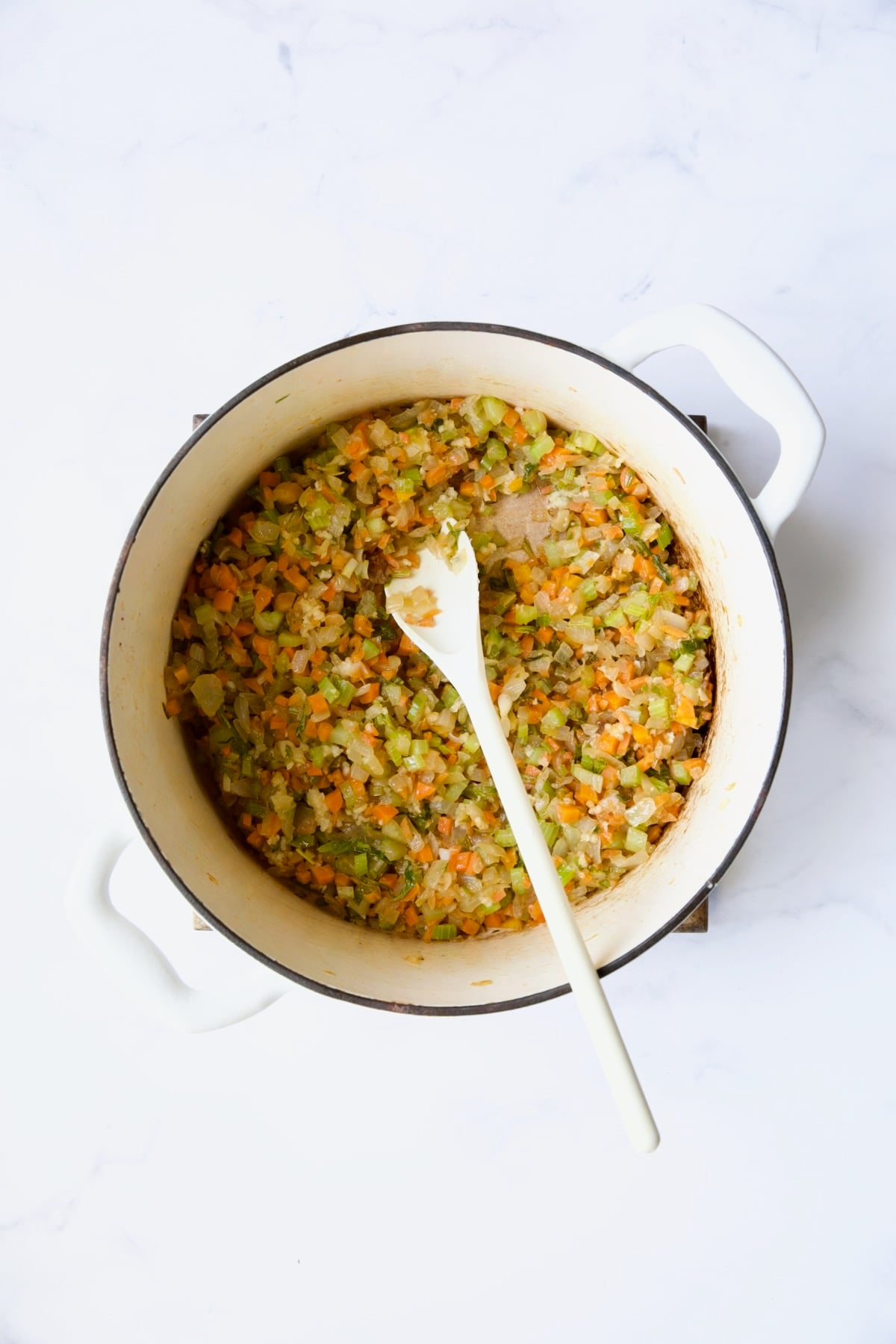 Cooked onions, celery and carrot in white Dutch oven being stirred with white spoon.