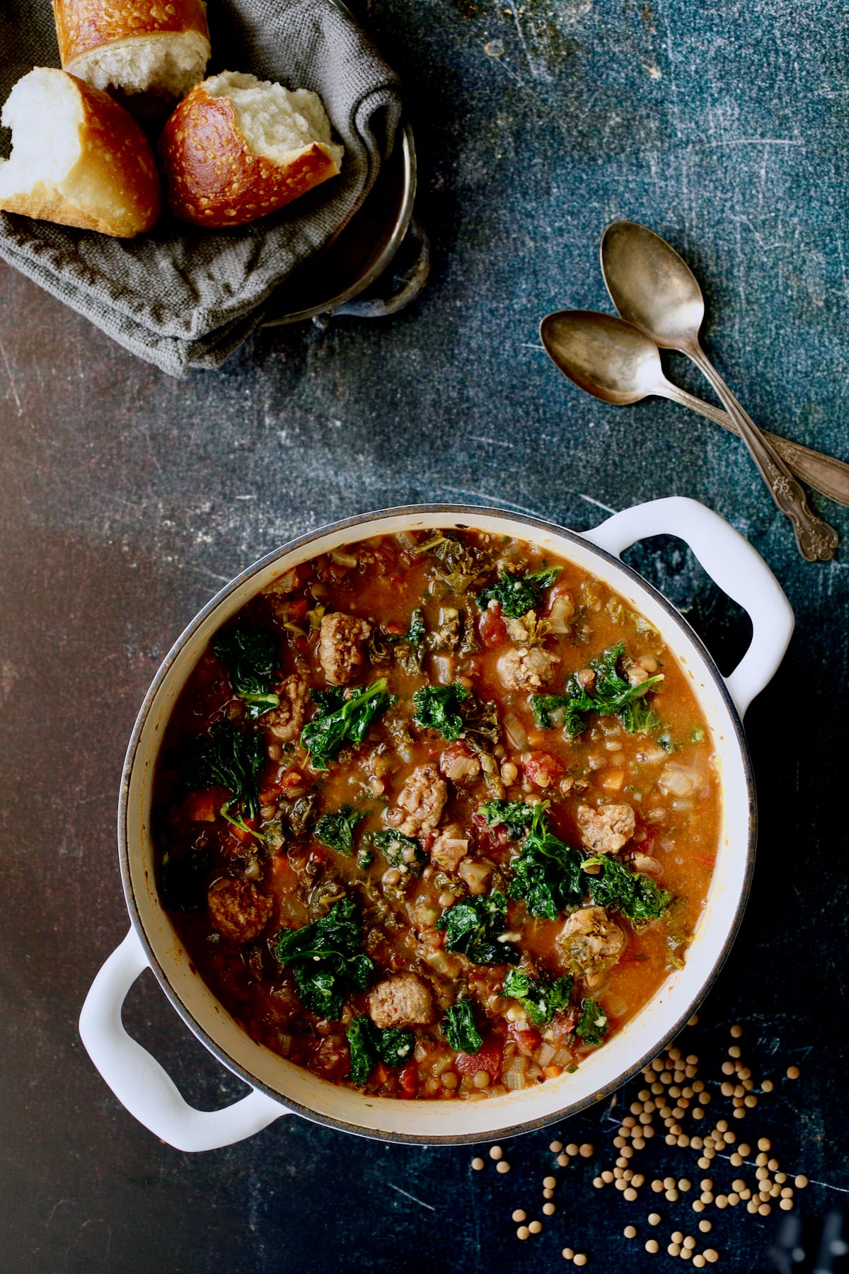 Sausage and Lentil Soup with Kale in white Dutch oven with bread, two spoons and lentils scattered about.