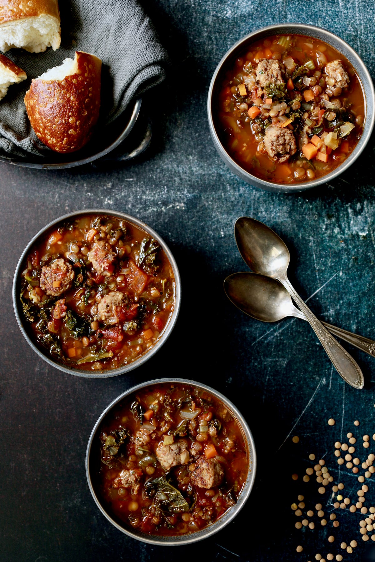 Three bowls of Sausage and Lentil Soup with Kale with bread on gray napkin.