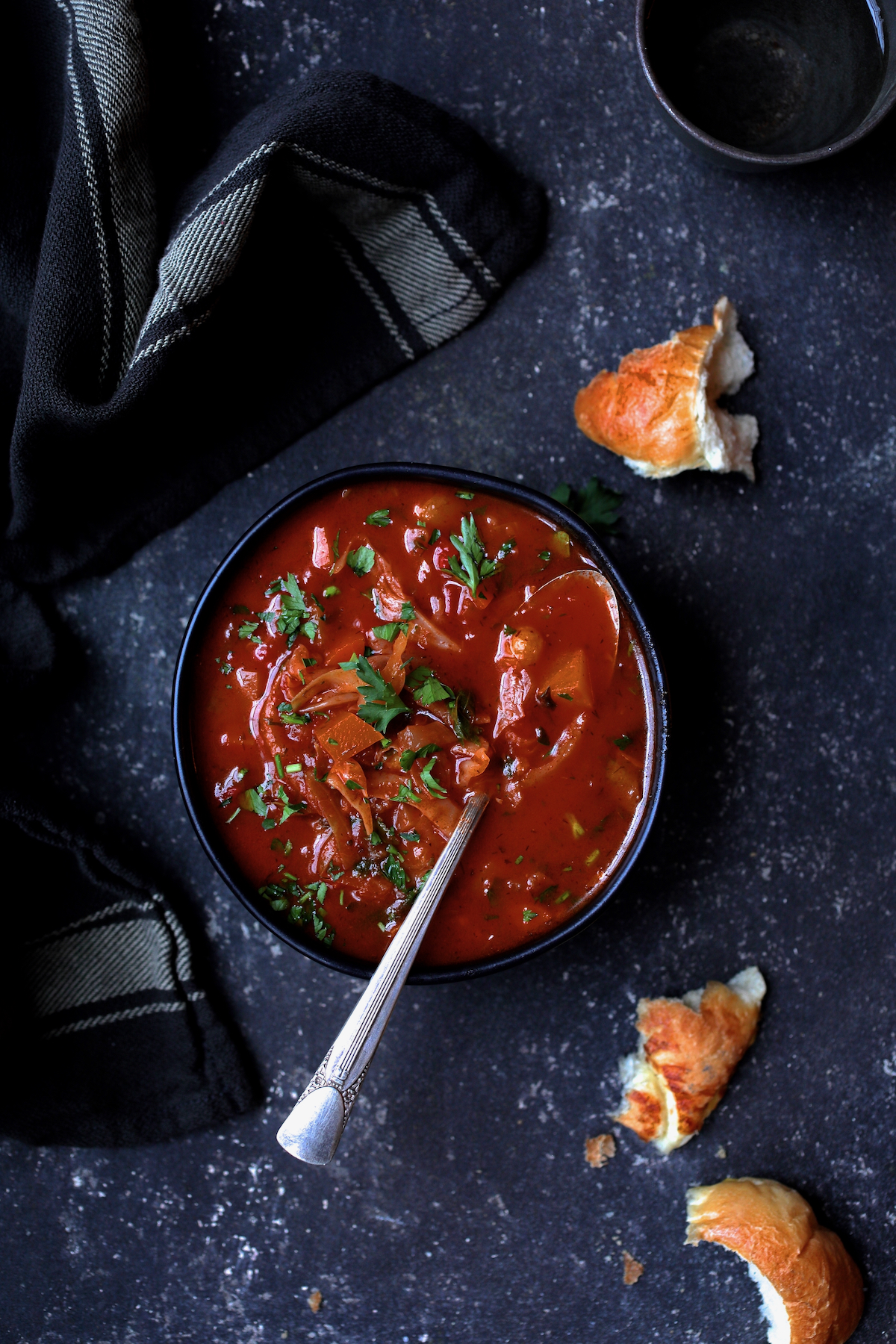 Rustic Cabbage and Root Vegetable Soup in black bowl garnished with parsley with pieces of bread scattered about.