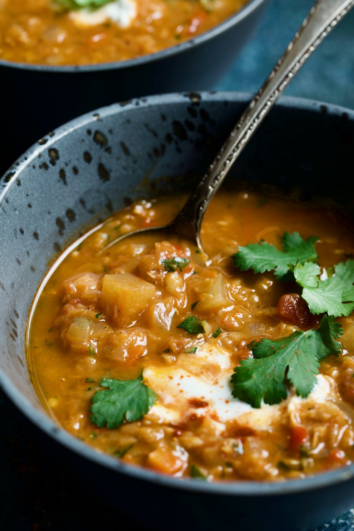 Photo of half bowl of Ethiopian Red Lentil Vegetable Soup in gray speckled bowl with spoon garnished with yogurt and cilantro.