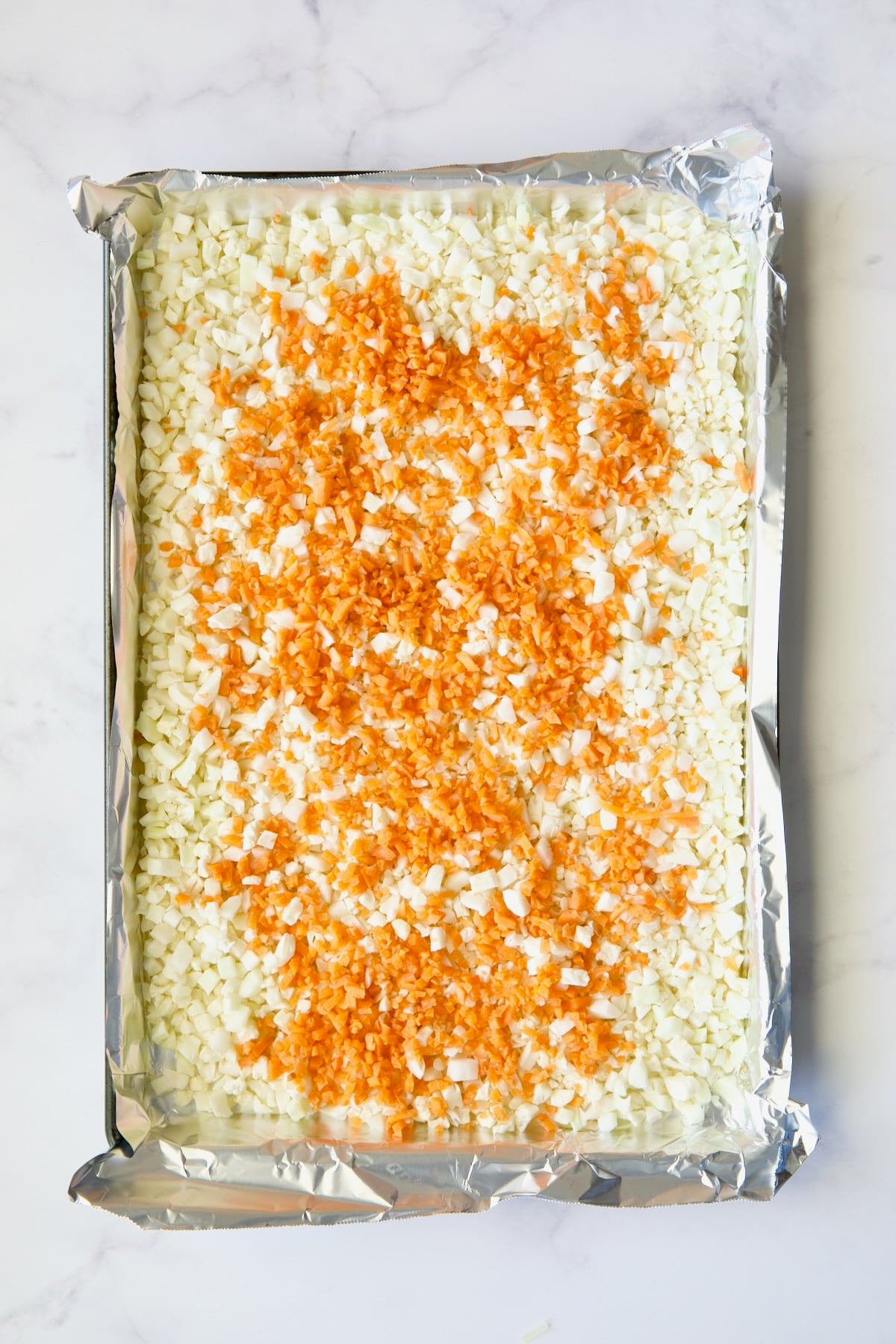 Cauliflower rice and diced carrot on aluminum foil-lined baking sheet before being roasted and before being stirred together.