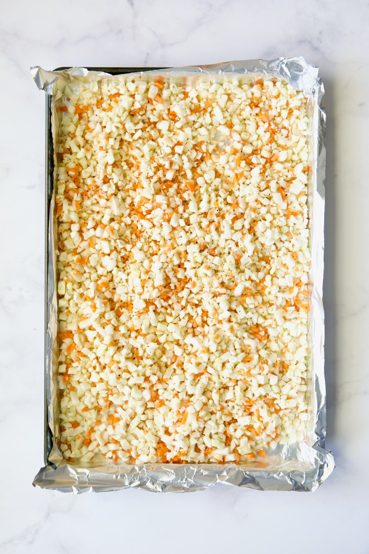 Cauliflower rice and diced carrot on aluminum foil-lined baking sheet before being roasted.