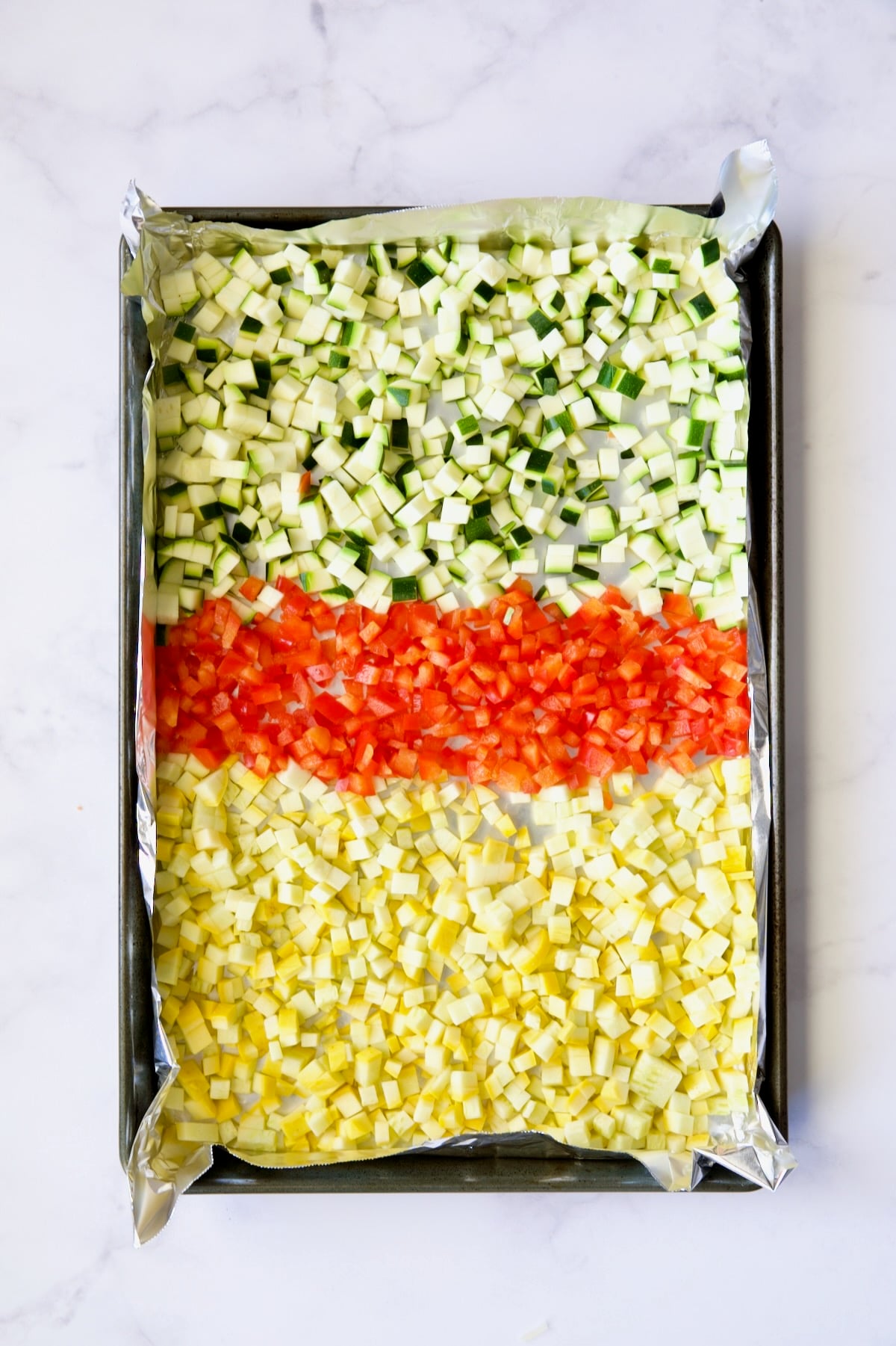 Diced zucchini, yellow squash and red bell pepper on aluminum foil-lined baking sheet before being roasted..