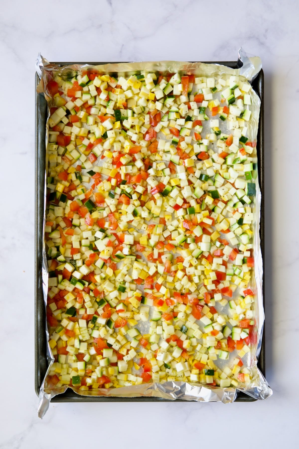 Diced zucchini, yellow squash and red bell pepper on aluminum foil-lined baking sheet before being roasted..