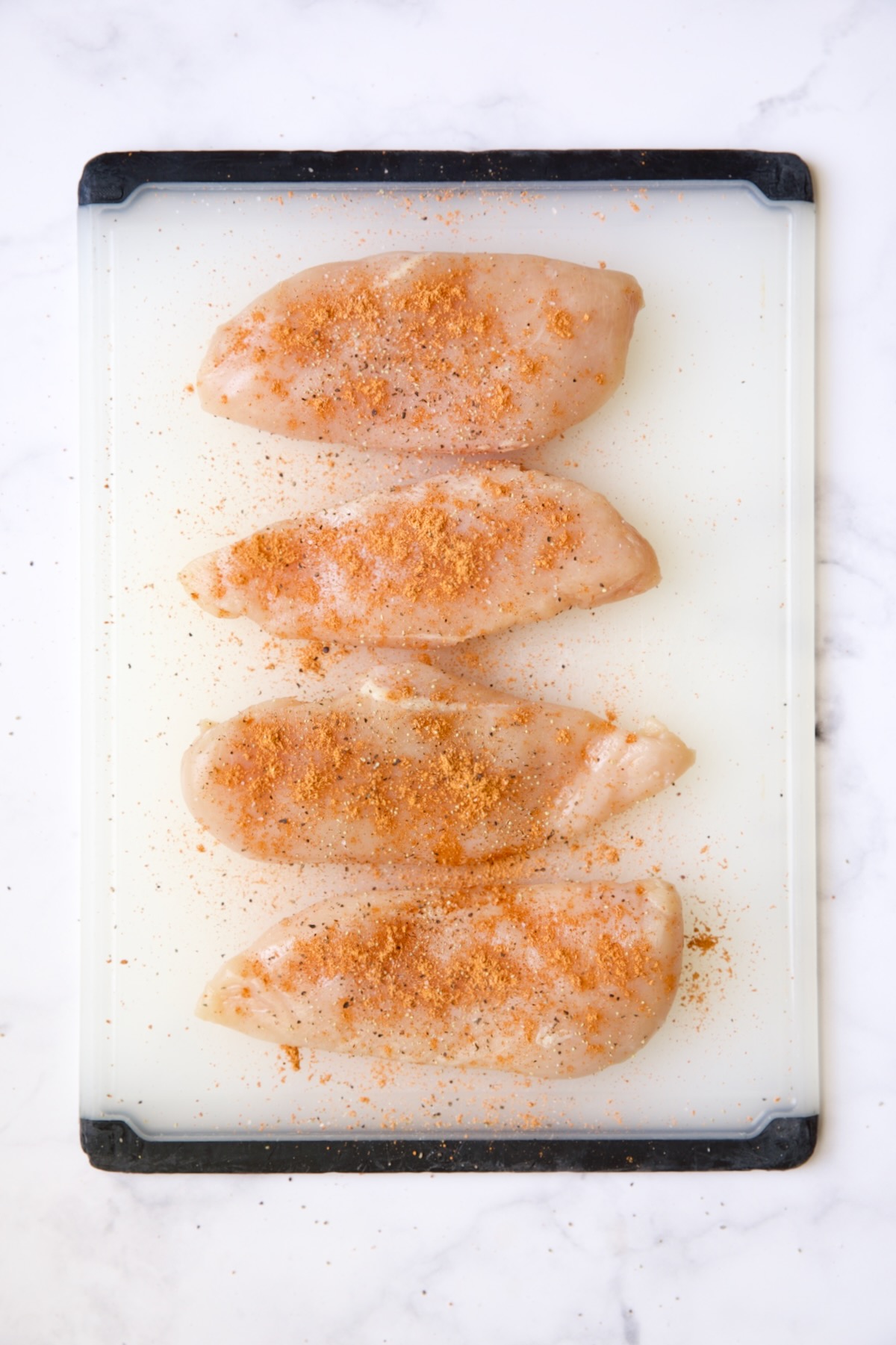 Four uncooked chicken breasts on white plastic cutting board sprinkled with taco seasoning.
