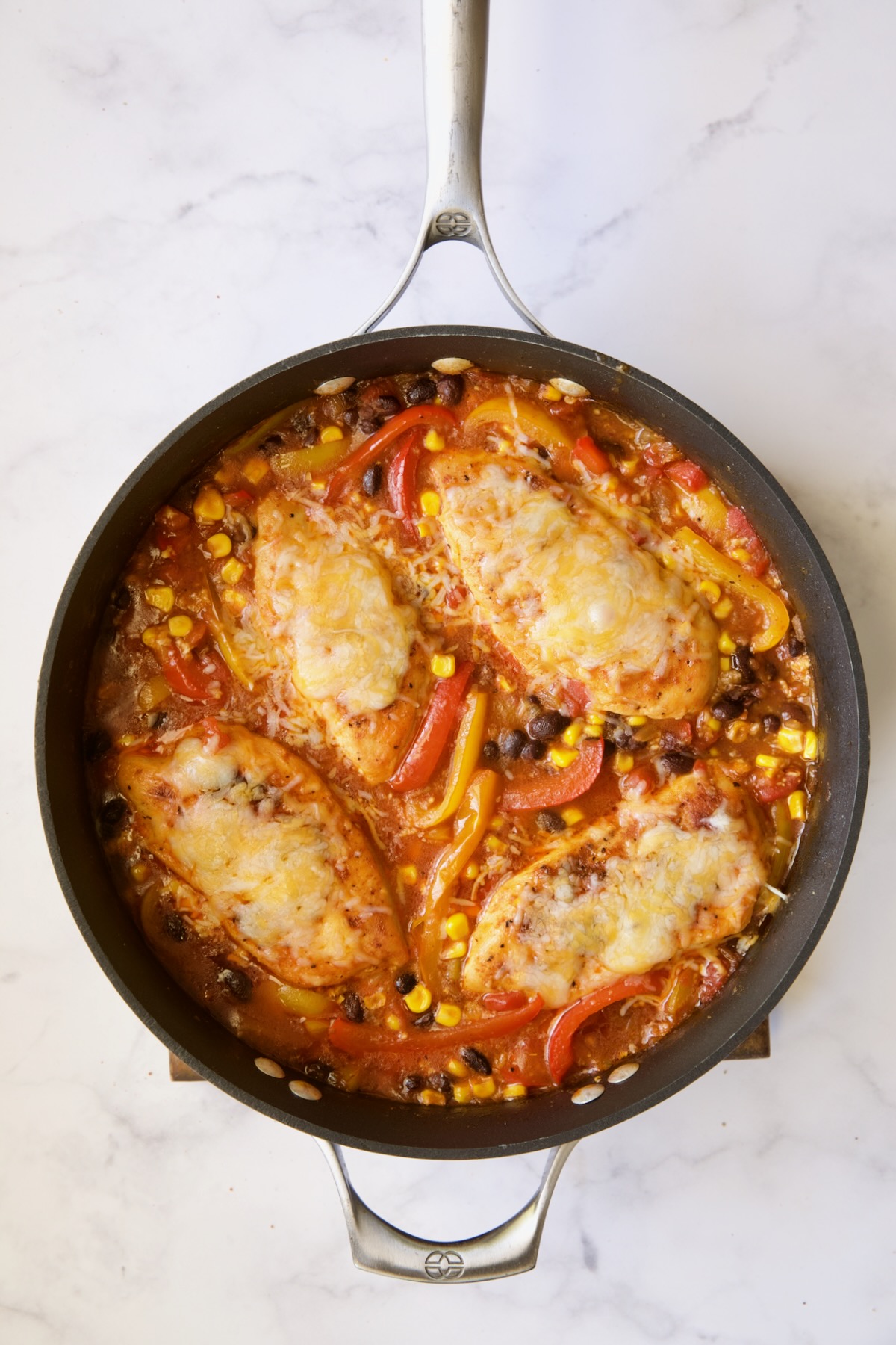 Four cheese-covered chicken breasts with other ingredients in skillet.
