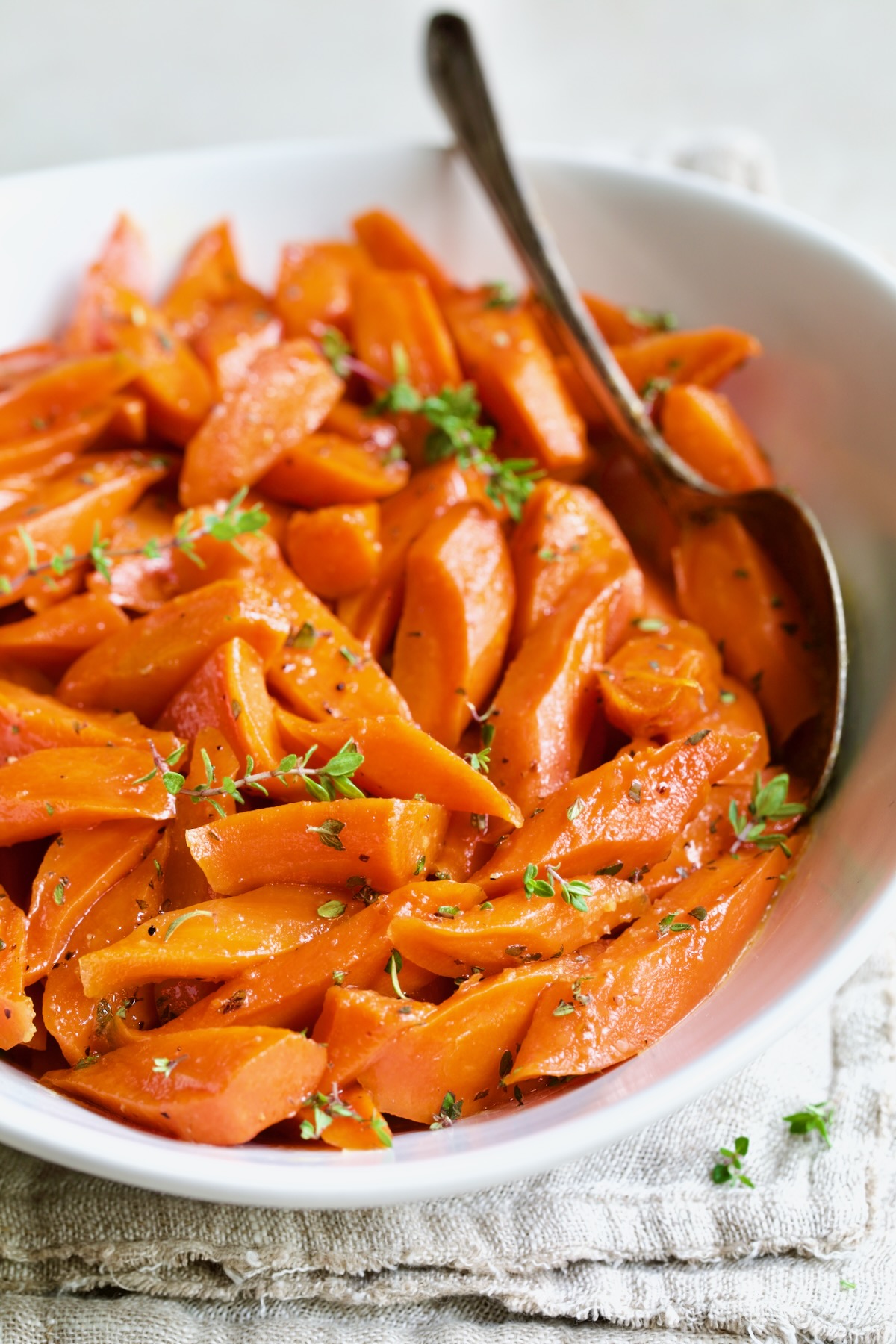 Bourbon-Glazed Carrots with Honey in white serving bowl with serving spoon garnished with fresh thyme.