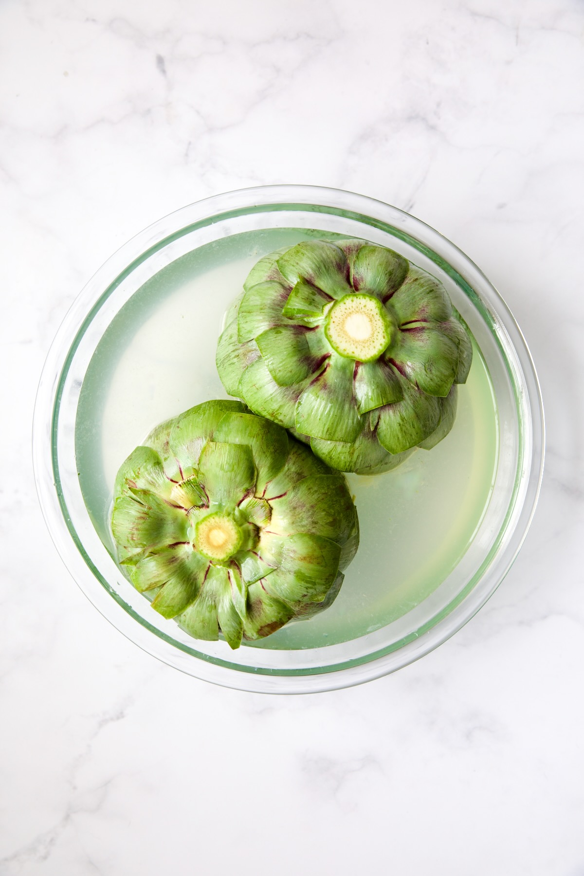 Two uncooked artichokes submerged in lemon water in glass bowl.