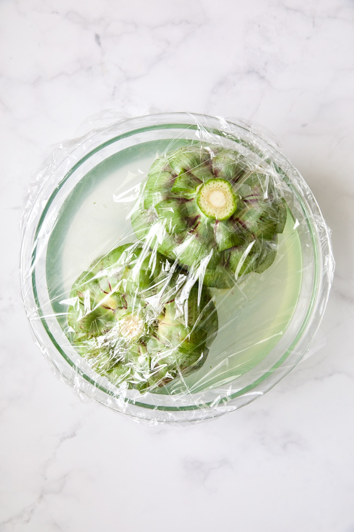 Two artichokes submerged in lemon water covered with plastic wrap.