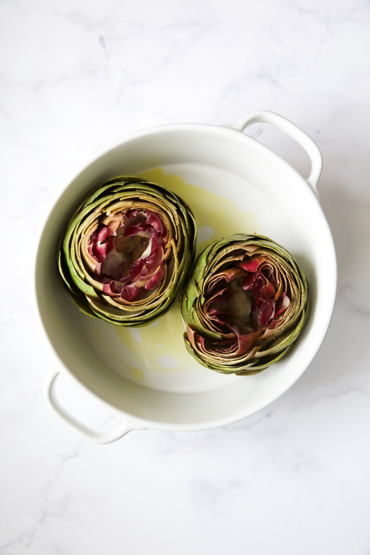 Two artichokes in white baking dish before being stuffed.