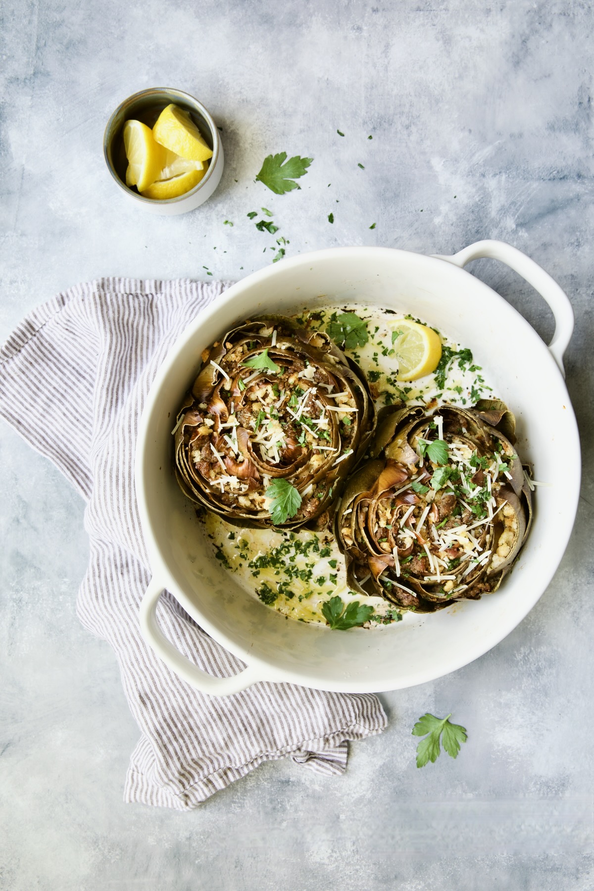 Two Italian-Stuffed Artichokes in white baking dish with lemon wedges and parsley.