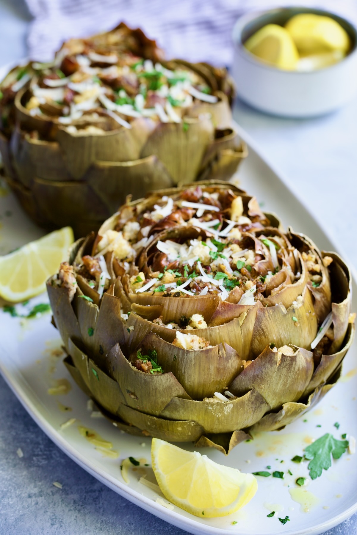 Two Italian-Stuffed Artichokes on white plates with lemon wedges.