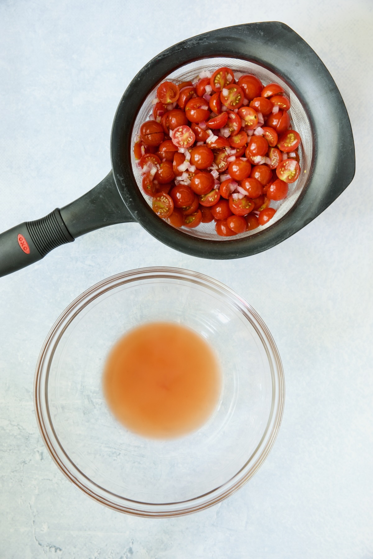 Cherry tomatoes and red onion in sieve and juices from in glass bowl.