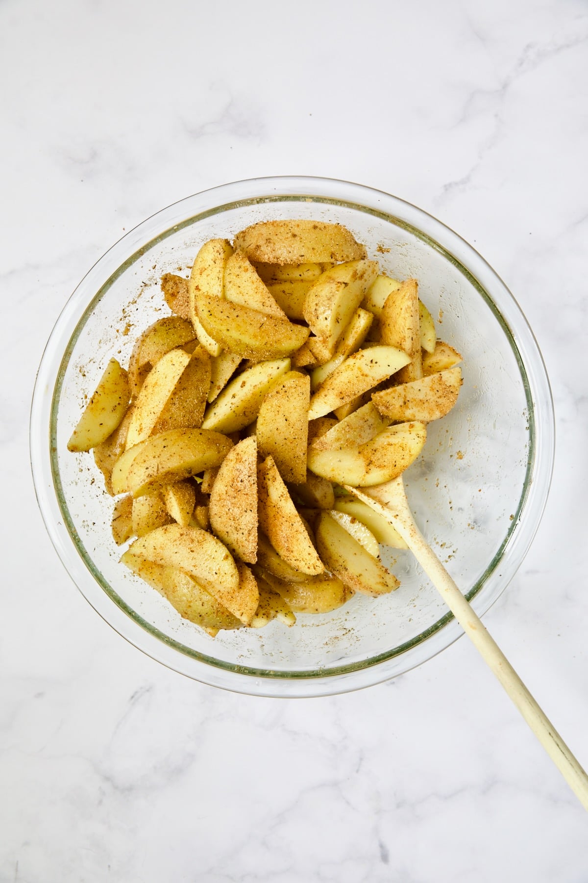 Seasoned Yukon Gold potato wedges in glass bowl being stirred with white spoon.
