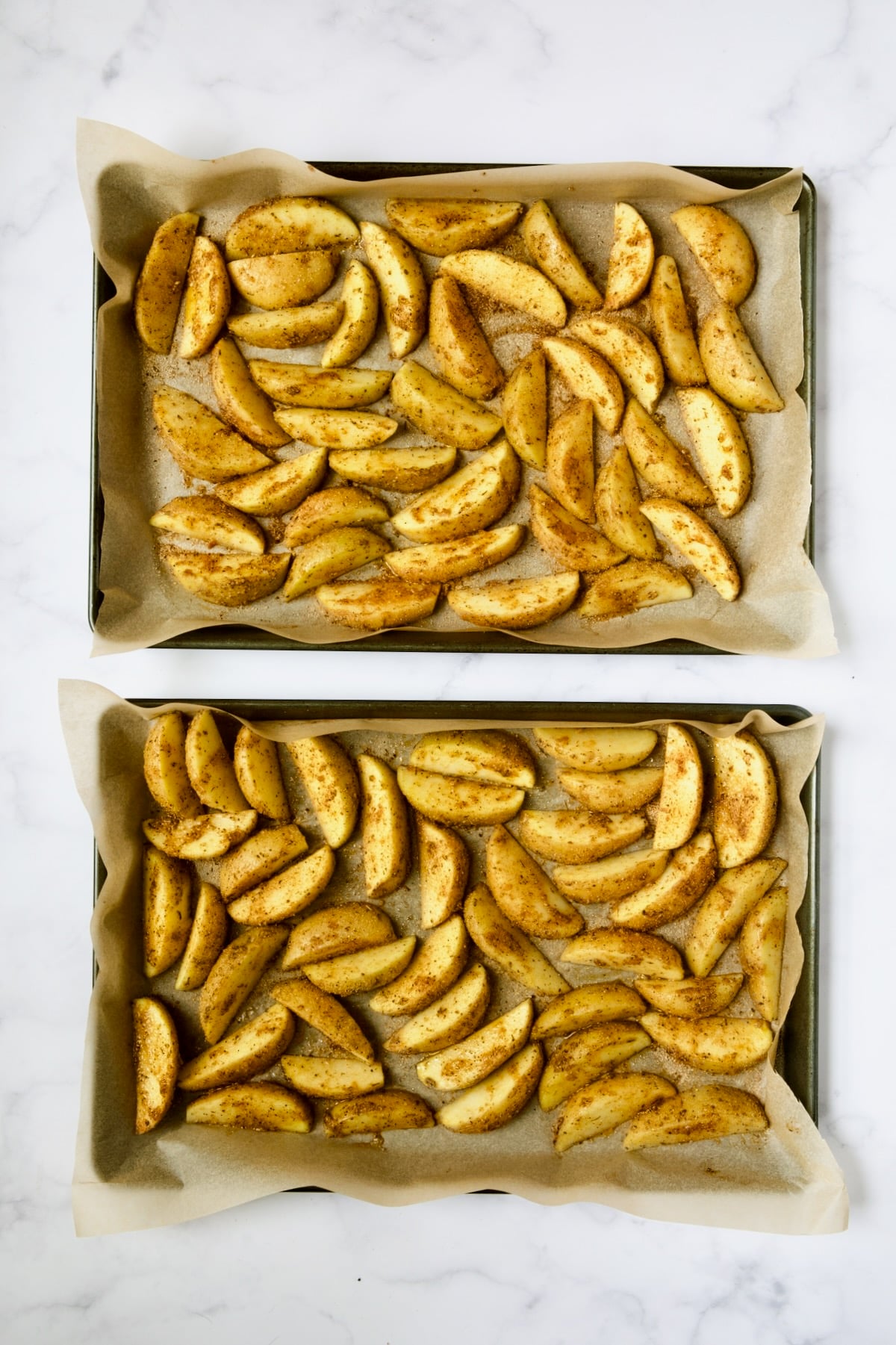 Two baking sheets lined with parchment paper with potato wedges before being roasted.