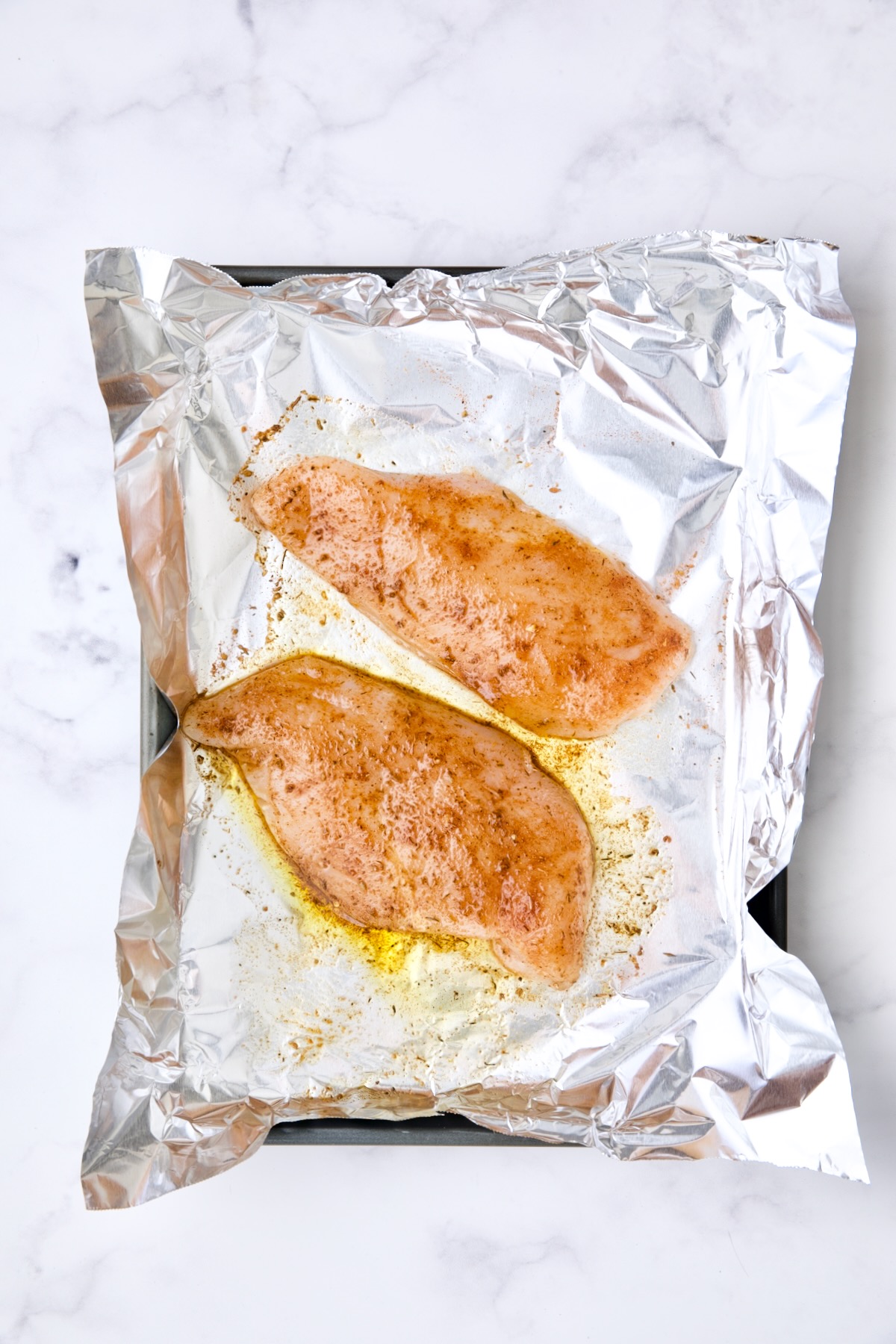 Uncooked chicken breasts on aluminum foil-lined rimmed baking sheets.