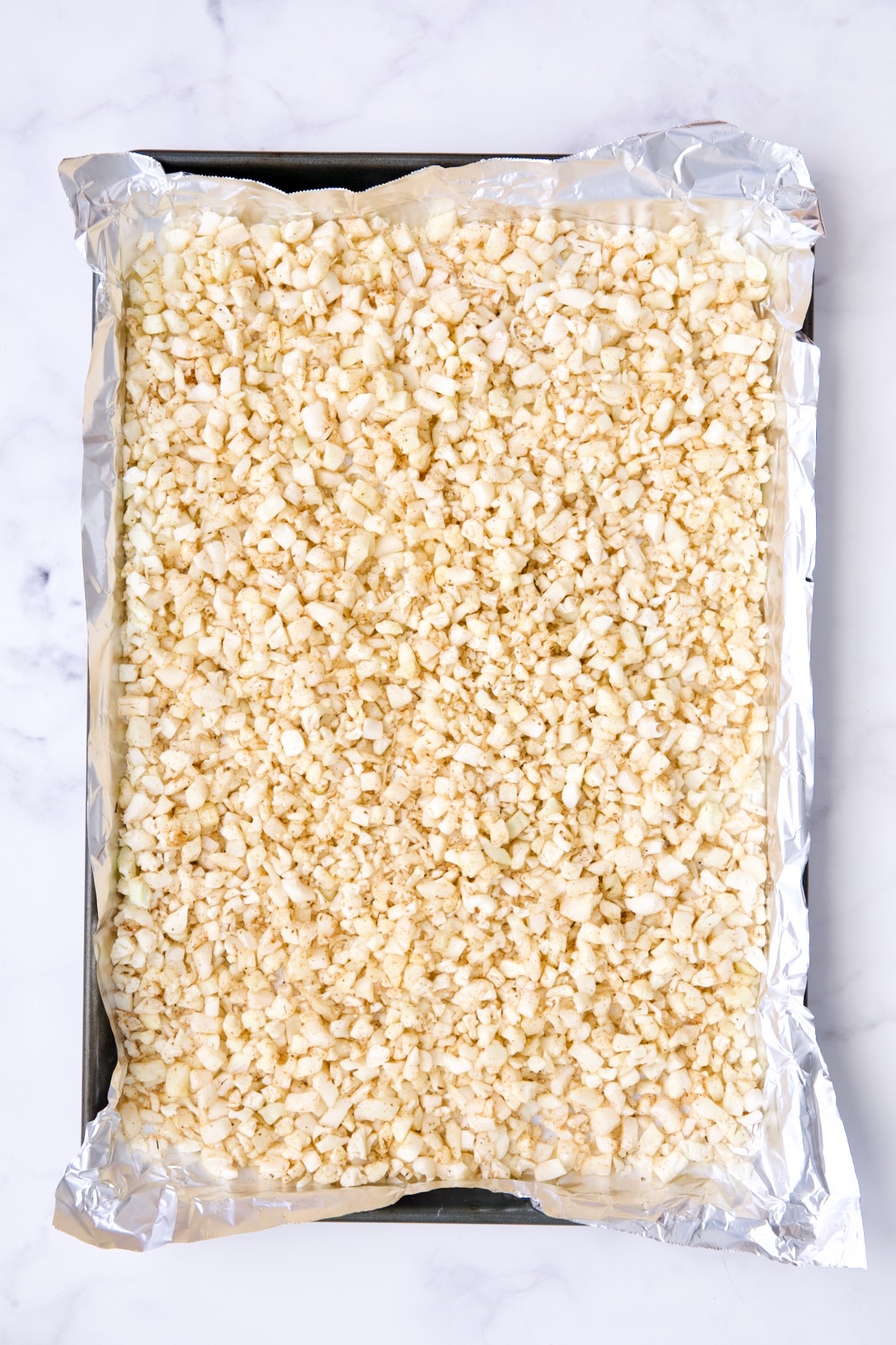 Uncooked riced cauliflower on aluminum foil-lined baking sheet.