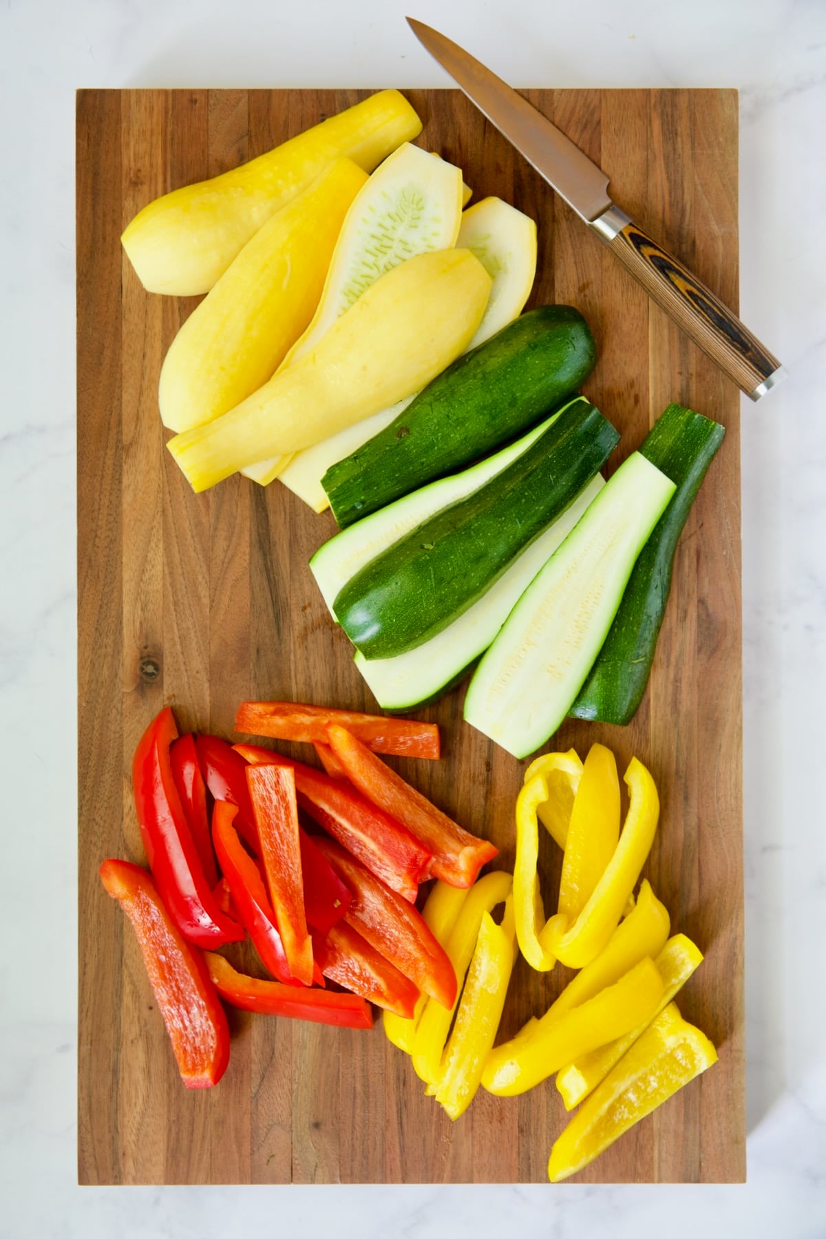 Cut zucchini, yellow squash and bell peppers on wooden cutting board with knife.