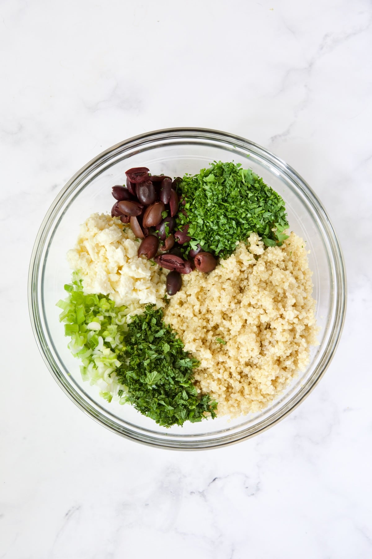Quinoa, olives, feta cheese, scallions, mint and parsley in glass bowl.