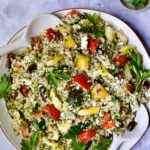 Mediterranean Quinoa Salad with Grilled Vegetables on round white platter with serving utensils garnished with herb sprigs.