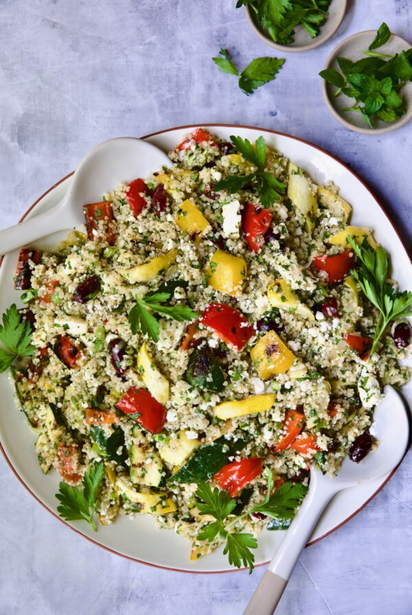 Mediterranean Quinoa Salad with Grilled Vegetables on round white platter with serving utensils garnished with herb sprigs.