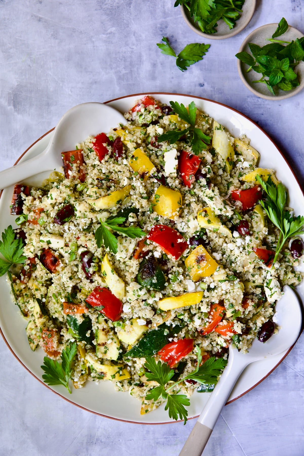 Mediterranean Quinoa Salad with Grilled Vegetables on round white platter with serving utensils garnished with herb sprigs.