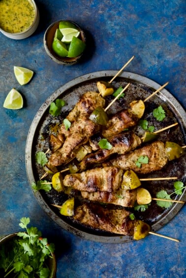 Mojo-Marinated Pork Skewers on metal tray with dipping sauce, lime wedges and cilantro leaves.