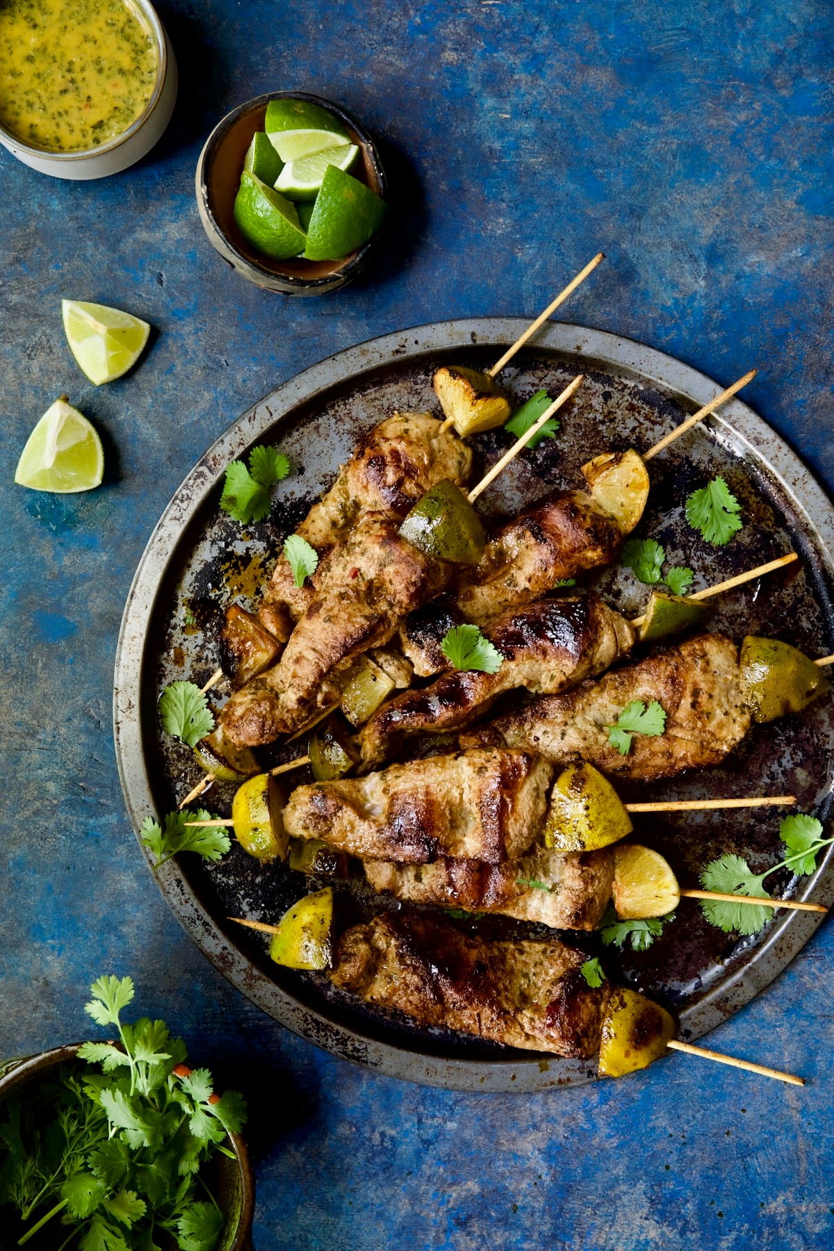 Mojo-Marinated Pork Skewers on metal tray with dipping sauce, lime wedges and cilantro leaves.